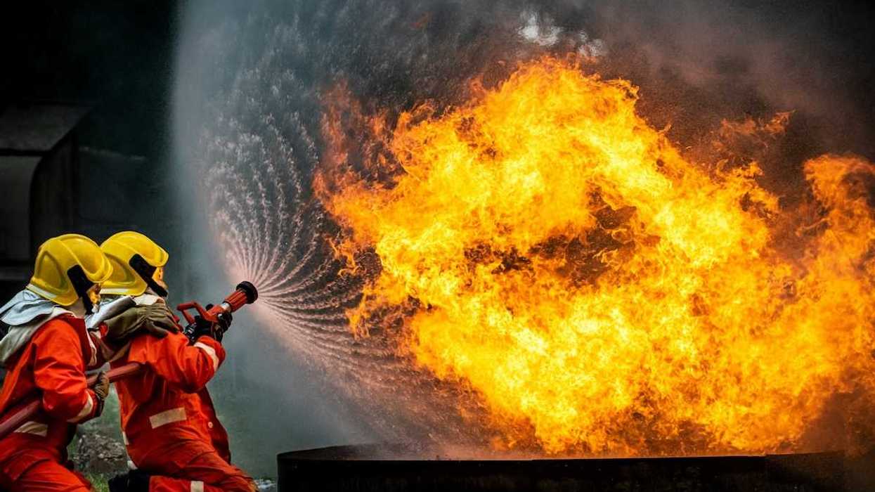 Firefighters dressed in red with yellow helmets training hose on chemical fire.