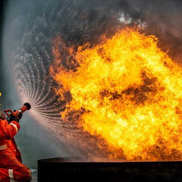 Firefighters dressed in red with yellow helmets training hose on chemical fire.