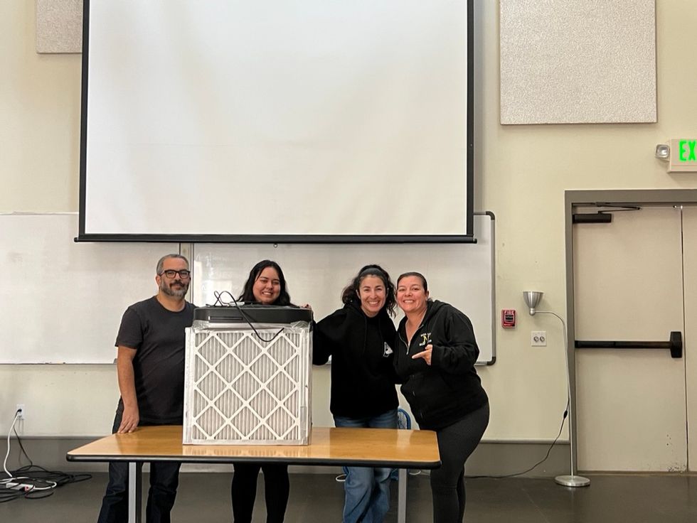 Four people pose with an indoor air filter in a classroom.