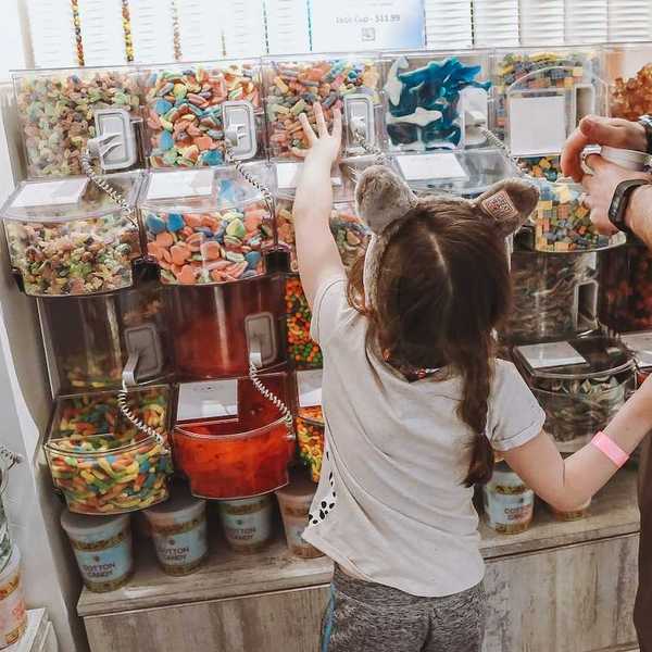 Girl standing with an adult, choosing candy at "bulk candy" counter in candy store.