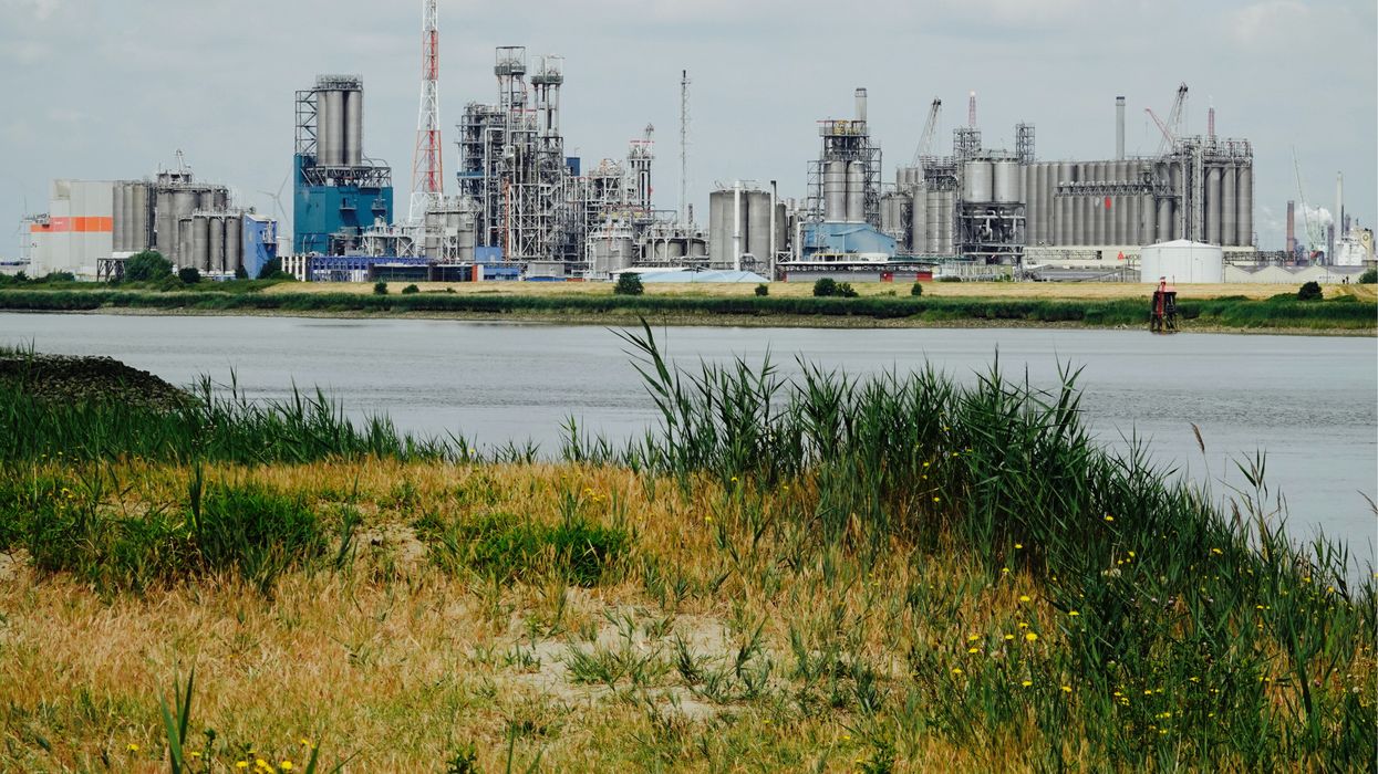 green grass field near body of water during daytime with industrial plants in the background.