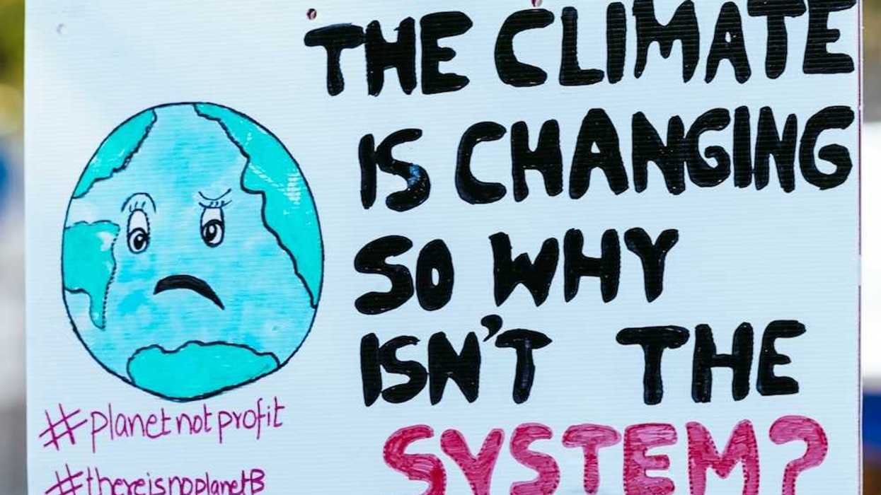 Hand holding handmade sign that reads "THE CLIMATE IS CHANGING SO WHY ISN'T THE SYSTEM?"