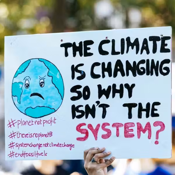 Hand holding handmade sign that reads "THE CLIMATE IS CHANGING SO WHY ISN'T THE SYSTEM?"