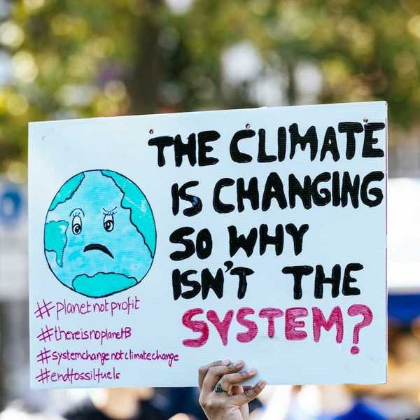 Hand holding sign: "THE CLIMATE IS CHANGING SO WHY ISN'T THE SYSTEM?"