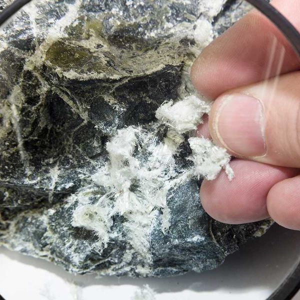 Hand with magnifying glass examines asbestos fibers in stone.