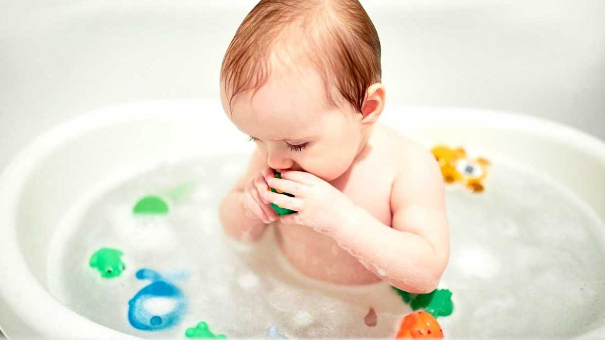 Infant in bathtub with plastic toy in mouth and surrounded by other plastic toys.