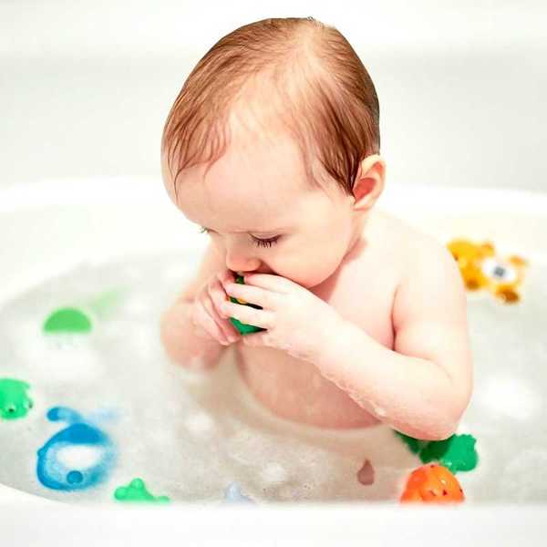 Infant in bathtub with plastic toy in mouth and surrounded by other plastic toys.