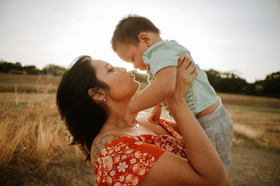 Latina woman in red and white floral dress kissing a baby in a golden field during daytime.