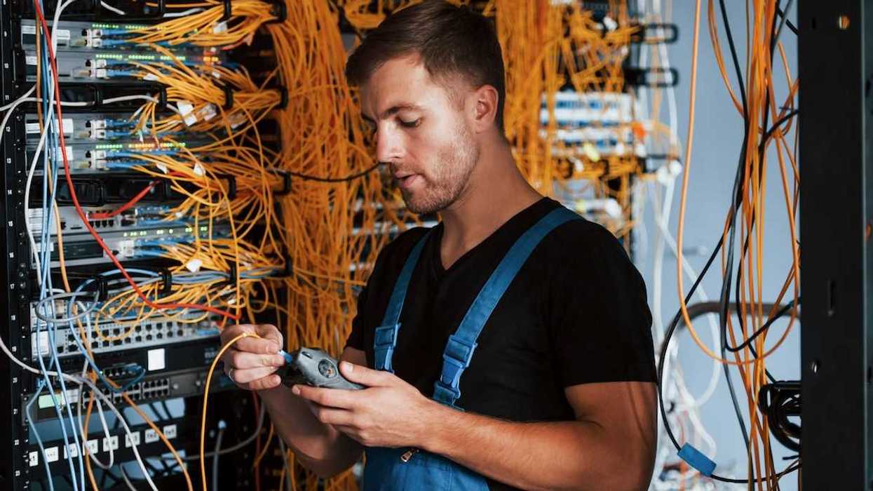 Male technician dressed in black t-shirt and blue bibbed overalls testing a rack of electronic gear