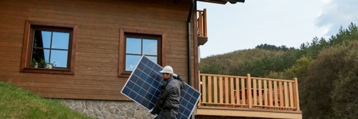 Man carrying a solar panel up a grass-covered slope near a home.