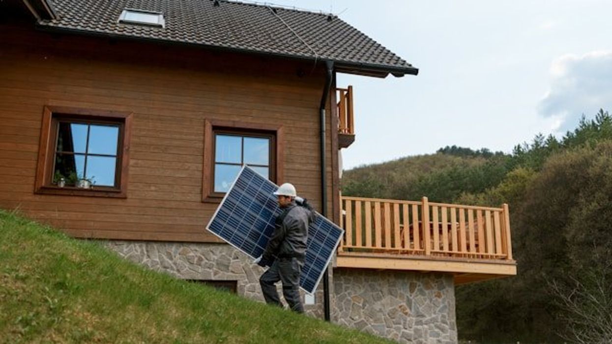 Man carrying a solar panel up a grass-covered slope near a home.