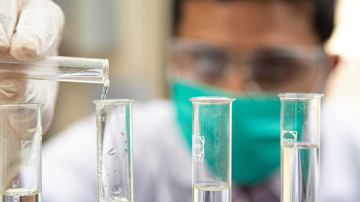 man in a surgical mask pouring clear liquid into clear glass test tubes.