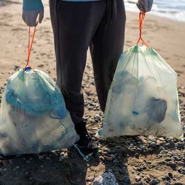 Man on a beach holding two large plastic bags filled with plastic trash