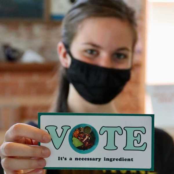 Masked woman holding small sign reading "VOTE It's a necessary ingredient"