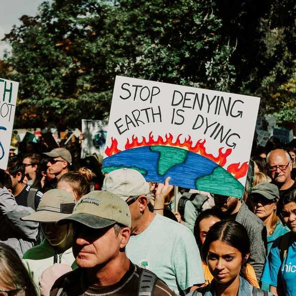 Mixed crowd taking part in climate protest prominently featuring a sign reading, "Stop Denying Earth is Dying"