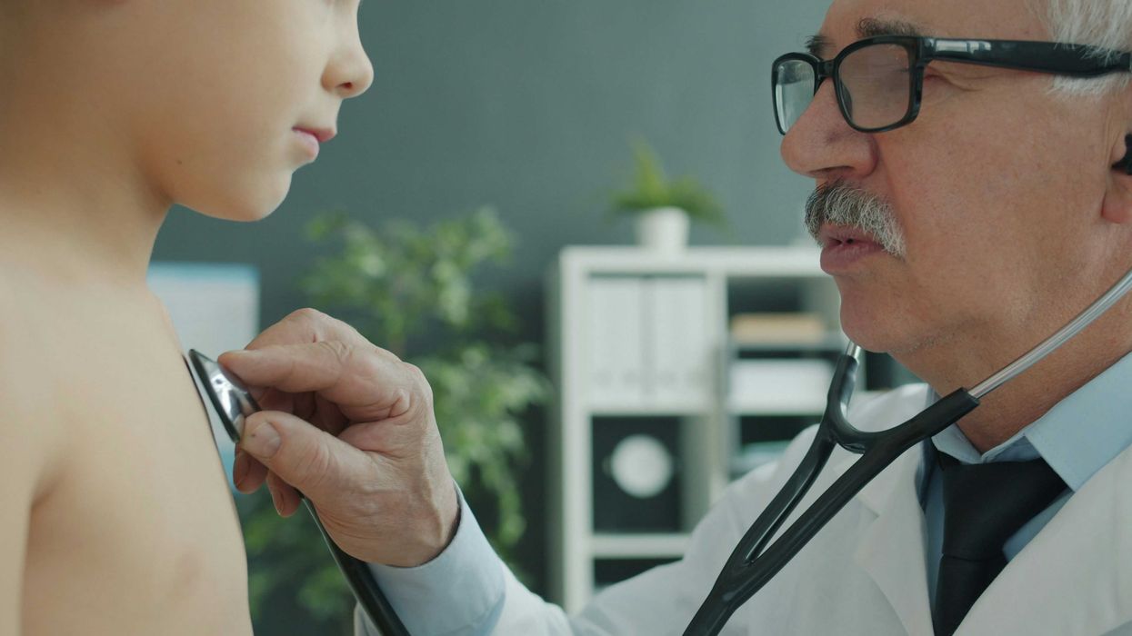 Older male doctor wearing glasses examines child's chest with stethoscope.