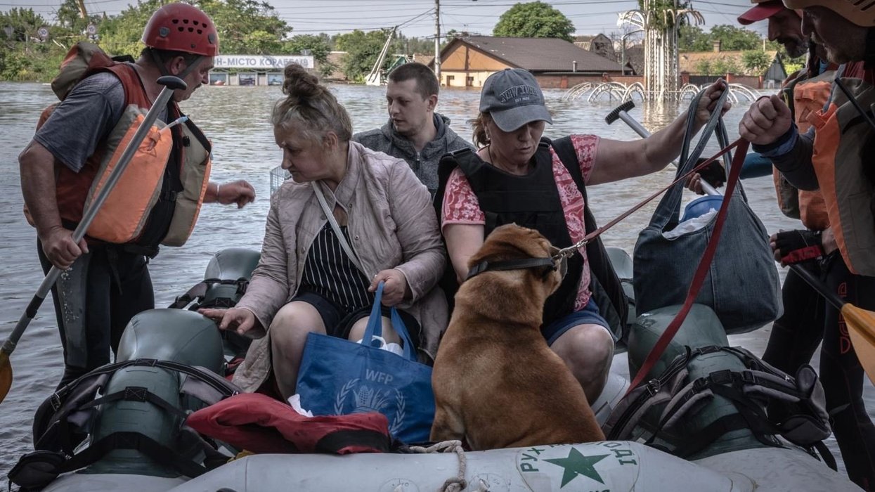 People and a dog in a rescue raft with floodwaters and flooded buildings in background.