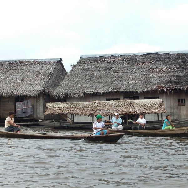 People in canoes on a river rowing past thatched roof wooden homes.