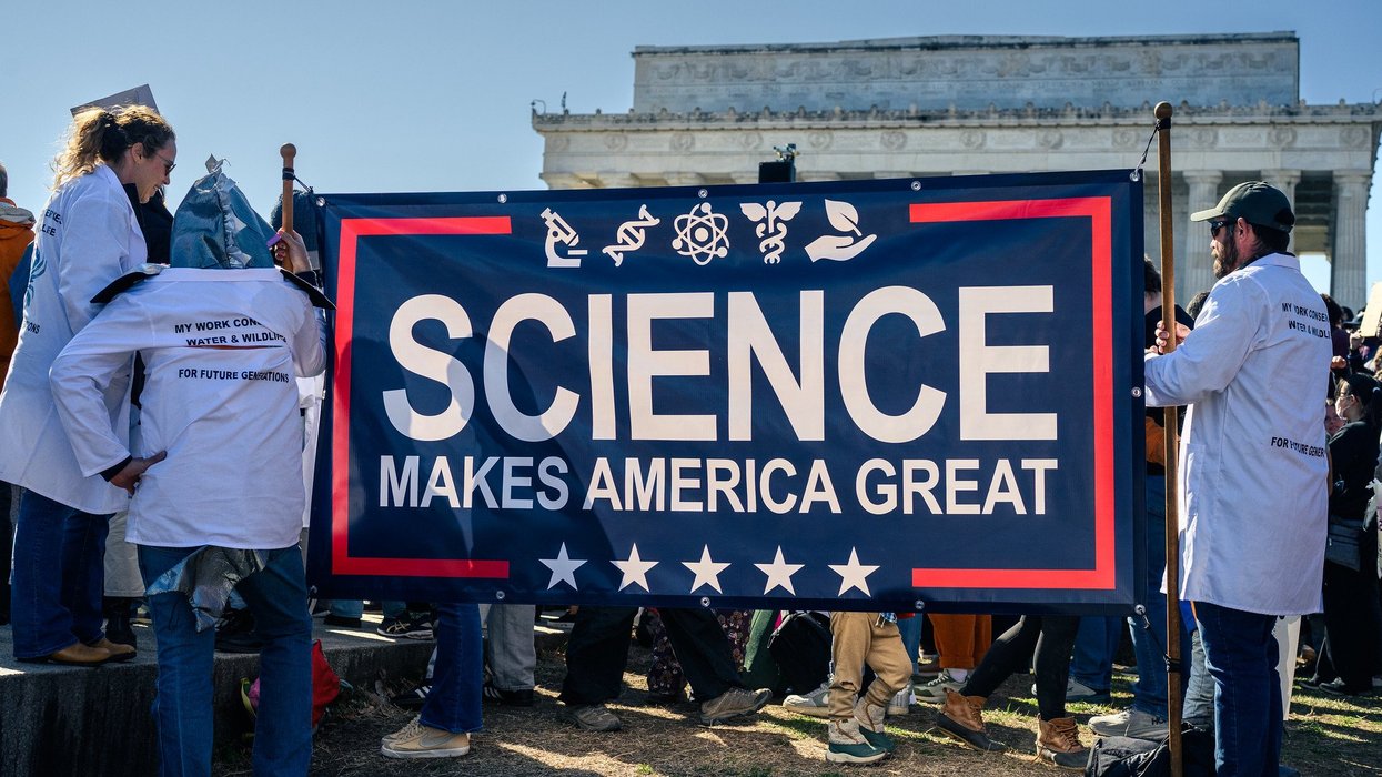 People in white lab coats stand outside with a large blue banner reading "science makes america great."