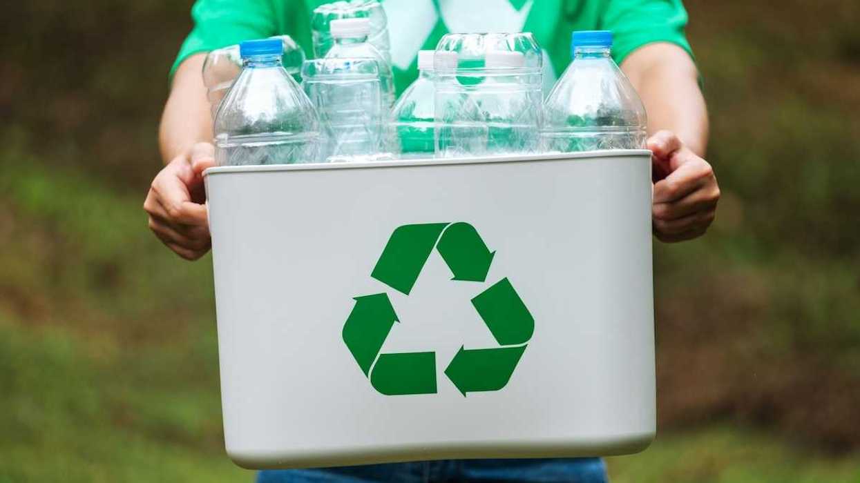 Person carrying a white plastic tote displaying a green recycling symbol full of plastic bottles