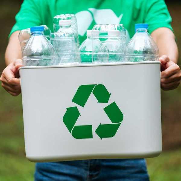 Person carrying a white plastic tote displaying a green recycling symbol full of plastic bottles