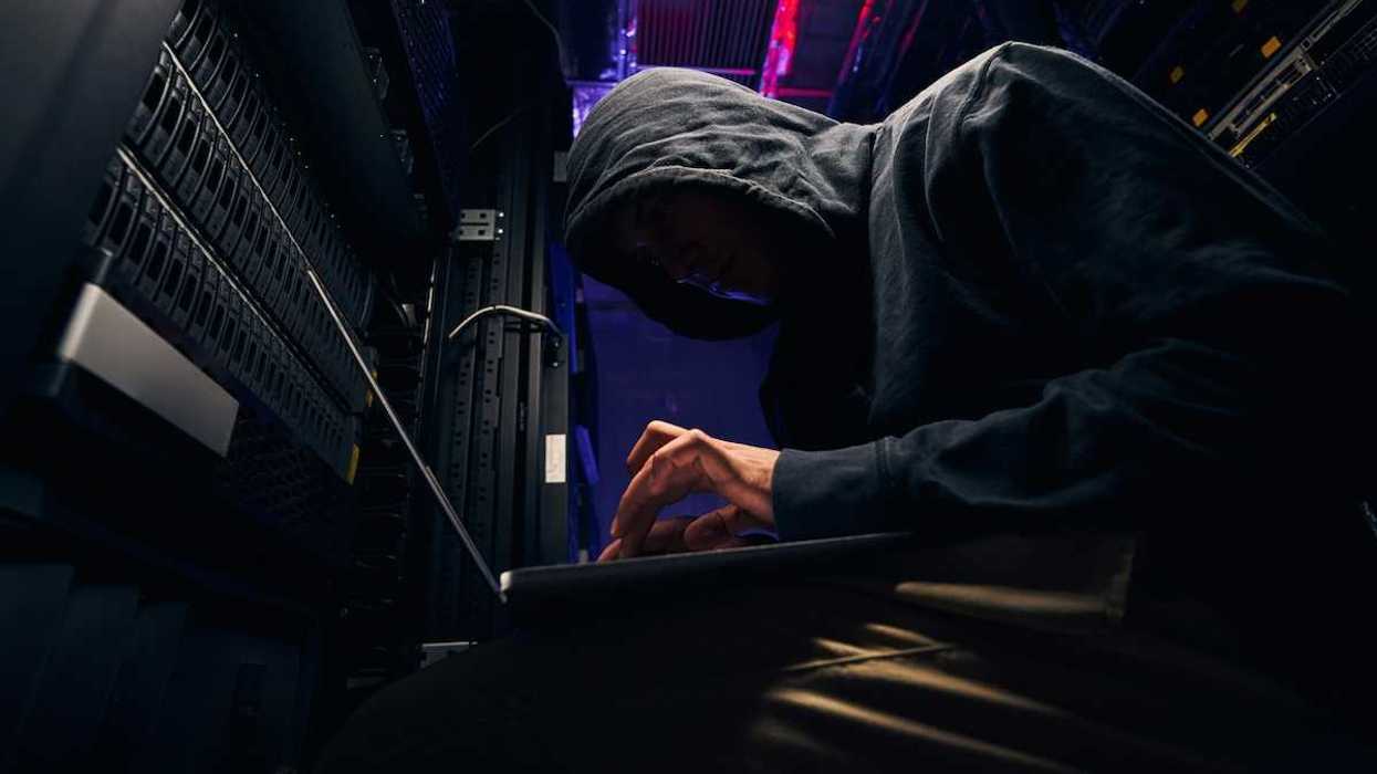 Person in black hoodie, hunched over a keyboard, personification of a shadowy computer hacker.