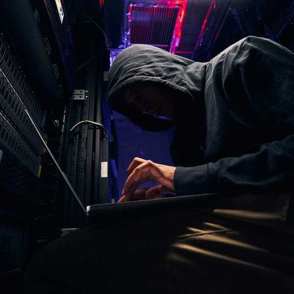 Person in black hoodie, hunched over a keyboard, personification of a shadowy computer hacker.