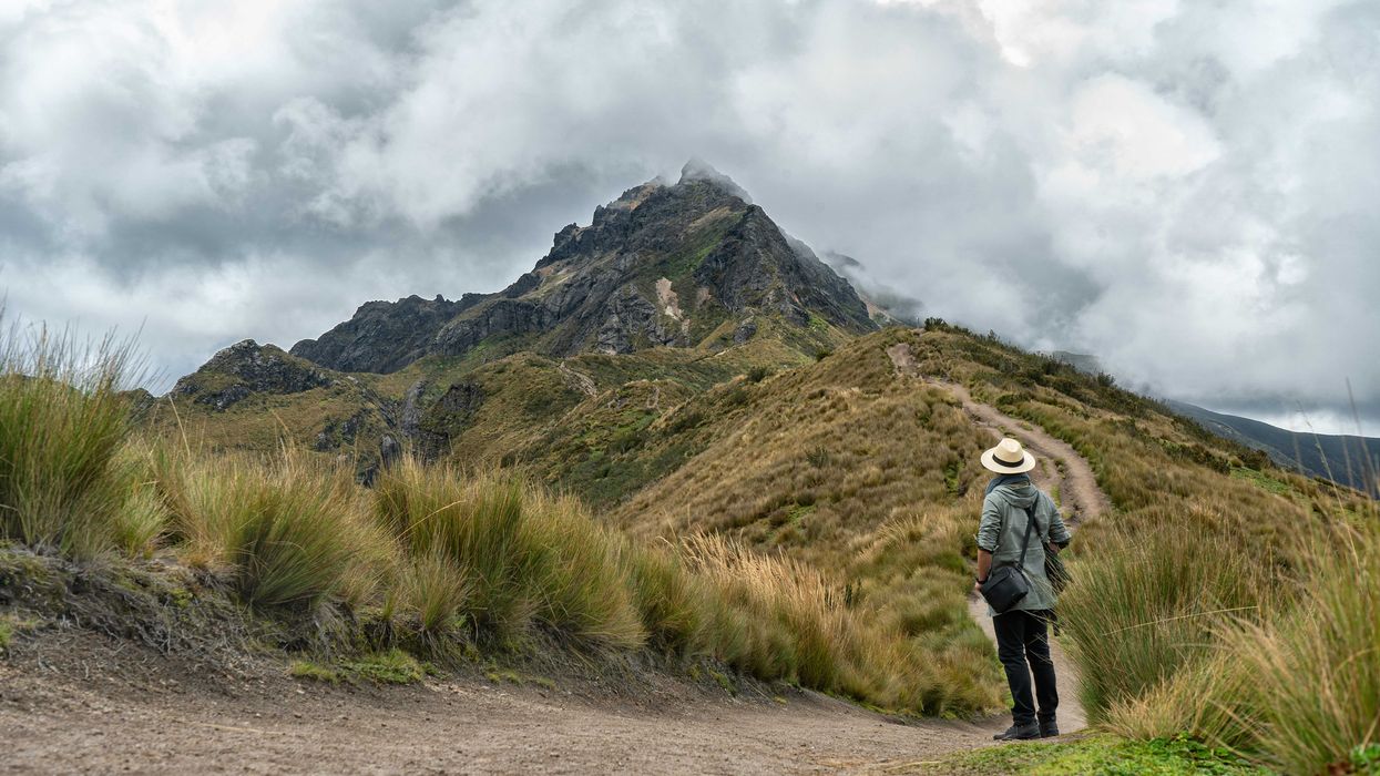 Person in gray jacket and black pants walking on dirt road in mist-covered mountains.