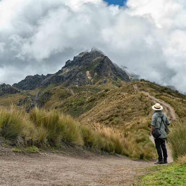 Person in gray jacket and black pants walking on dirt road in mist-covered mountains.