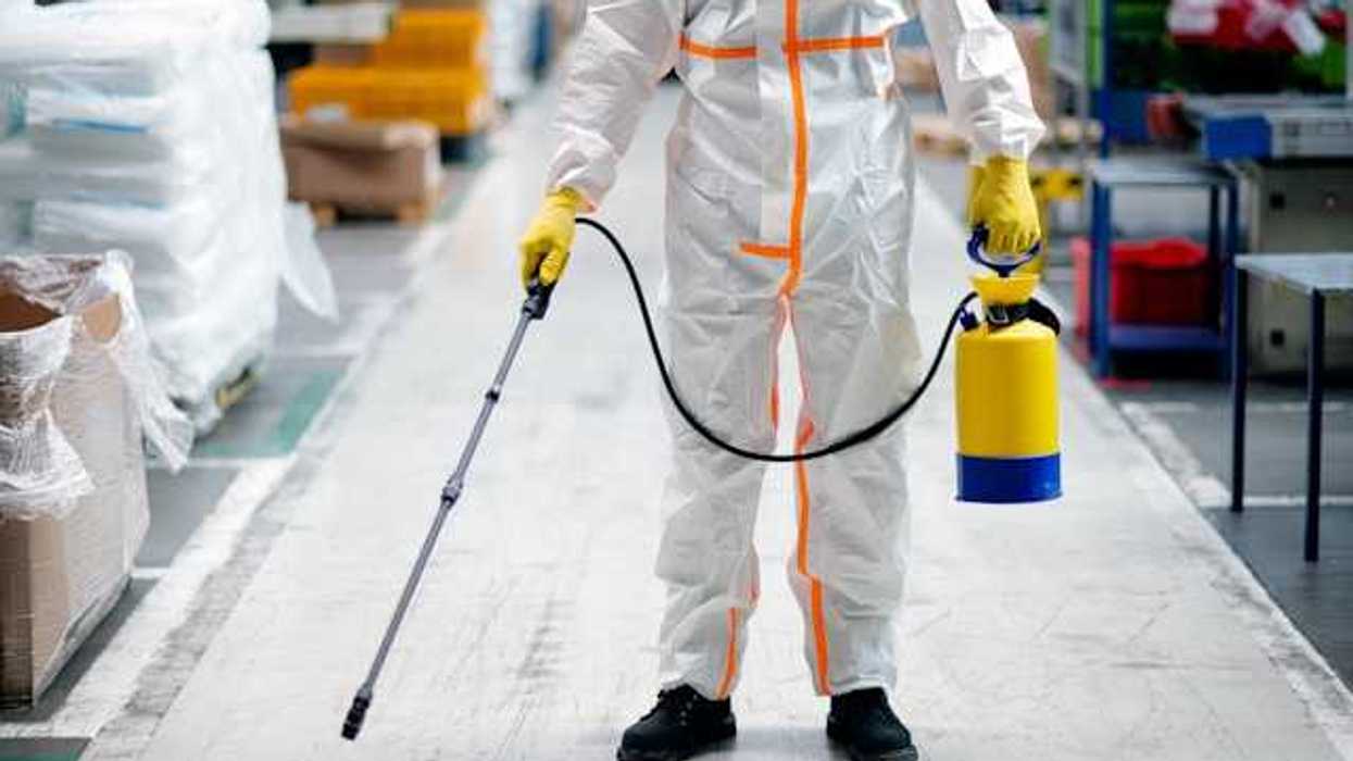 Person in white protective clothing with pesticide container and long-armed spray wand in hand