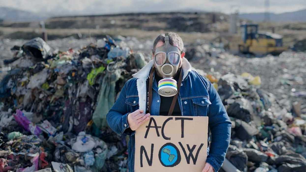 Person standing in an active landfill, wearing respirator and holding a cardboard sign reading "ACT NOW"