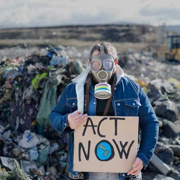 Person standing in an active landfill, wearing respirator and holding a cardboard sign reading "ACT NOW"