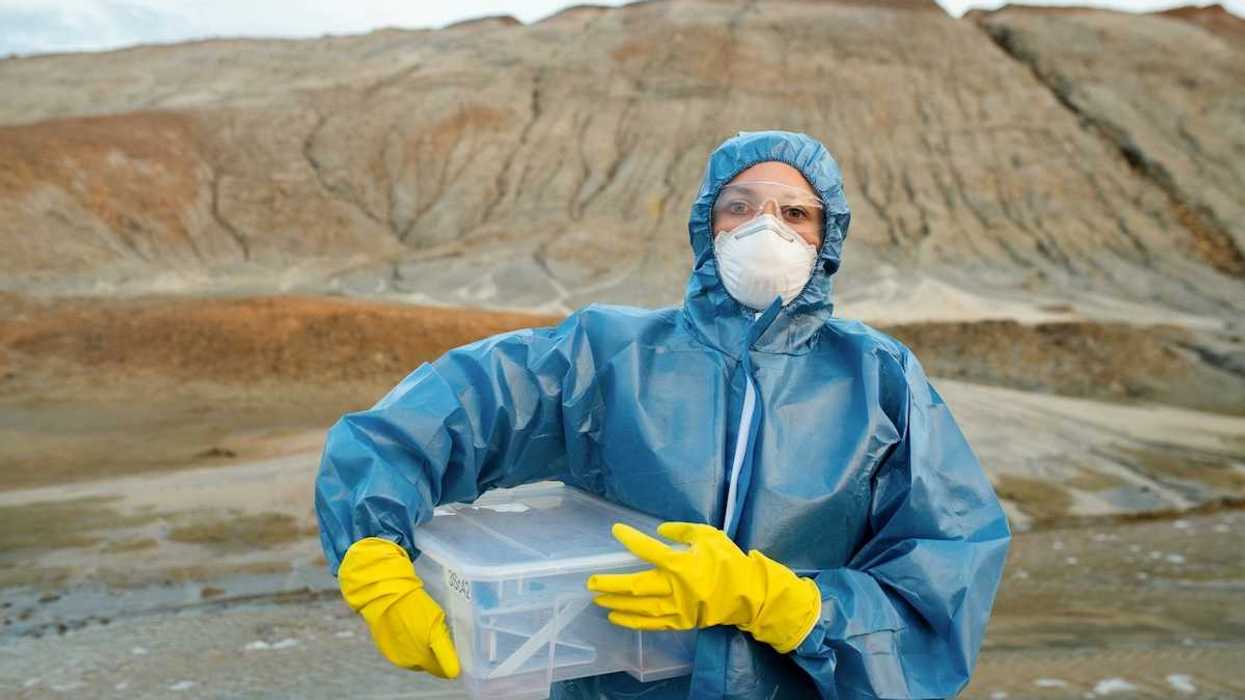 Person wearing blue protective suit, white particle mask, clear goggles and yellow gloves standing in front of large earthen mound.