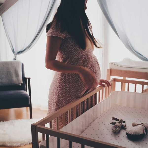 pregnant woman in a dress standing beside brown wooden crib.