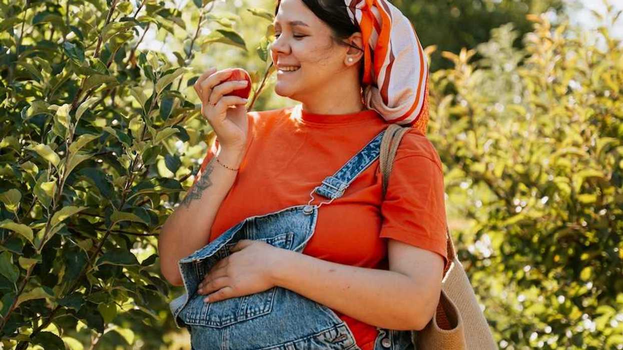 Pregnant woman in denim overalls and orange jersey eating an apple fresh off the tree