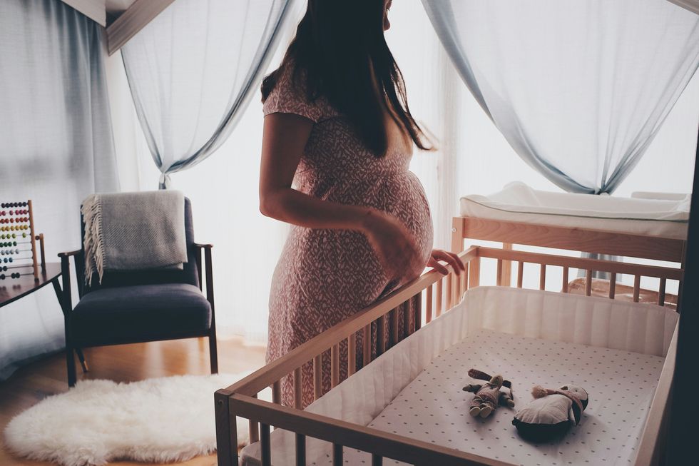 Pregnant woman in short-sleeved long dress standing beside brown wooden crib with window in background.