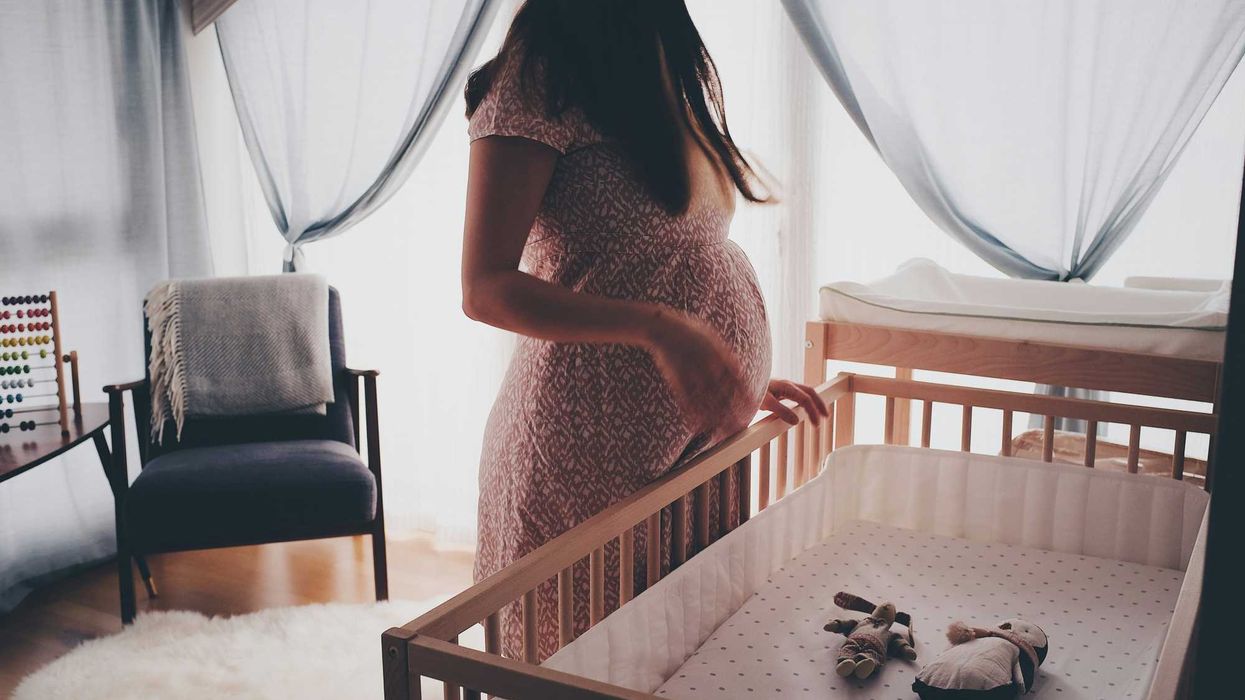 Pregnant woman in white lace sleeveless dress standing beside brown wooden crib.