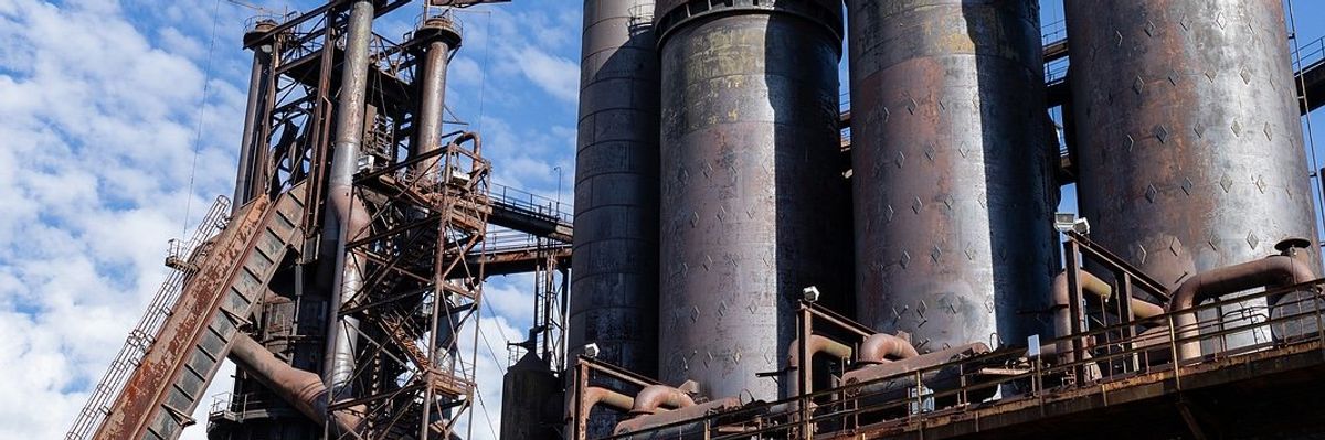 Rusting abandoned steel mill, blast furnaces and smokestacks atop a concrete wall.