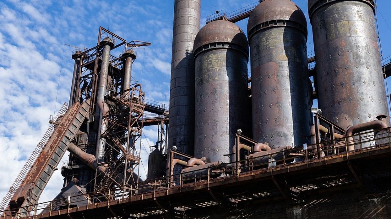 Rusting abandoned steel mill, blast furnaces and smokestacks atop a concrete wall.