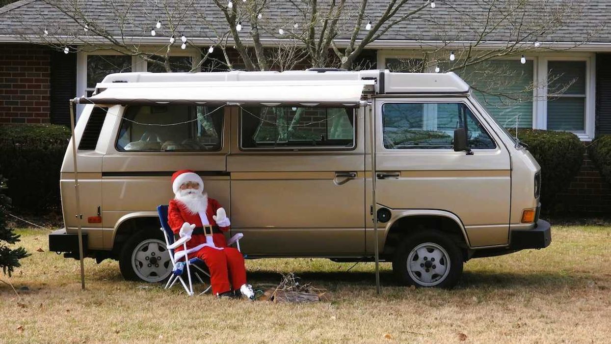 'Santa Claus' sitting in lawn chair adjacent to a camper van on snowless lawn