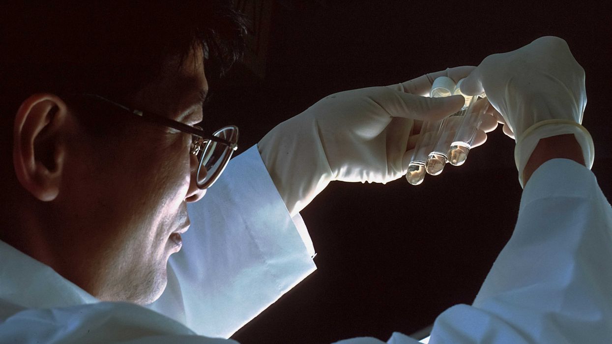 scientist in white lab coat and gloves holding test tubes up to the light.