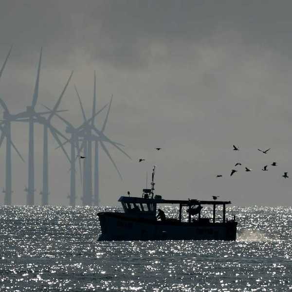 Small fishing boat trailed by sea birds with wind turbines in the background.