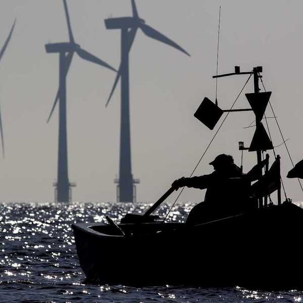 Small one-man fishing boat silhouetted at sunrise with sustainable energy wind turbines in the background.