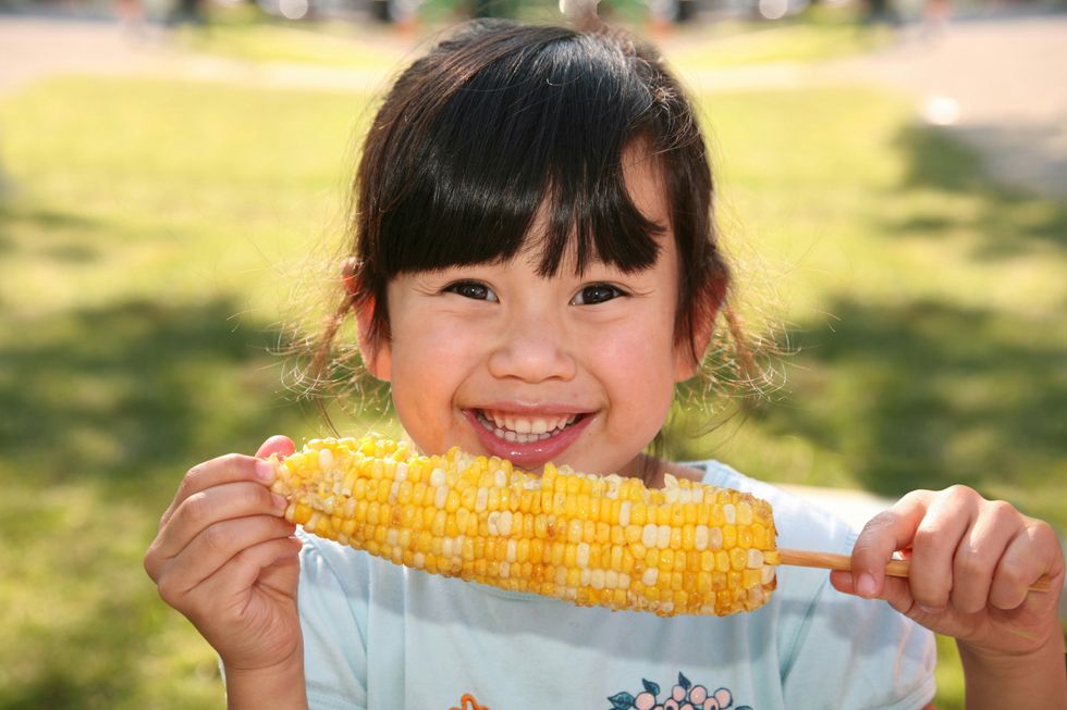 smiling girl holding corn on the cob in front of her face.