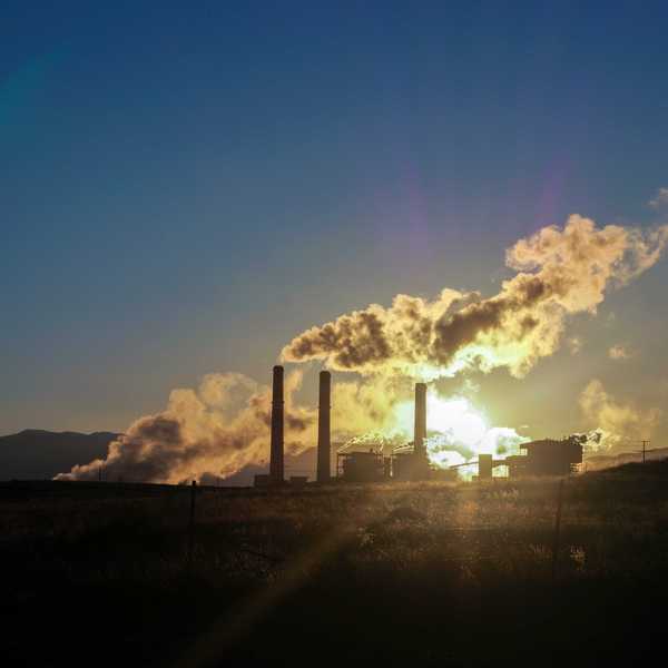 Smoke from smokestacks blows in the wind with sun shining in background.