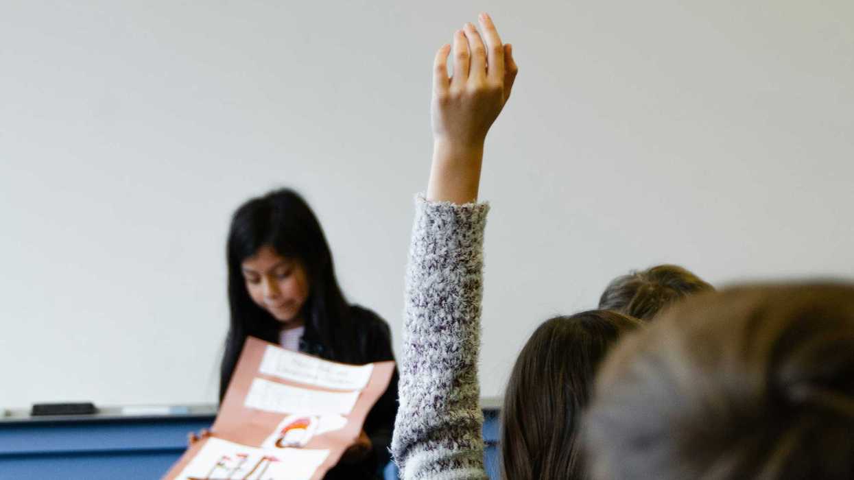 student raising hand in classroom while another student presents a poster.