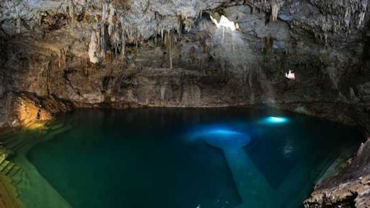 Sunlight streams into a cave cenote, illuminating clear blue water beneath dramatic limestone formations.