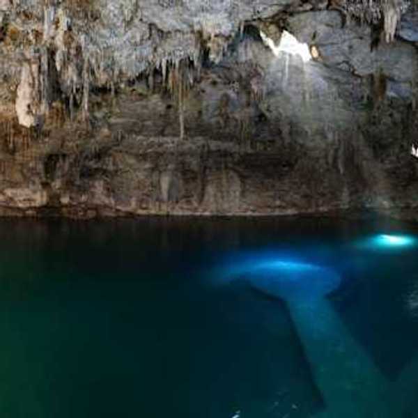 Sunlight streams into a cave cenote, illuminating clear blue water beneath dramatic limestone formations.