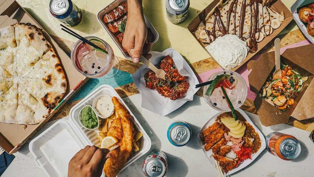 Table of takeout food featuring a variety of packaging and two hands indicating consumption in progress