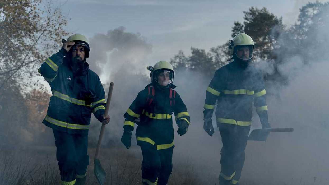 Three firefighters in turnout gear — woman in the center, flanked by two men carrying shovels in smoky air